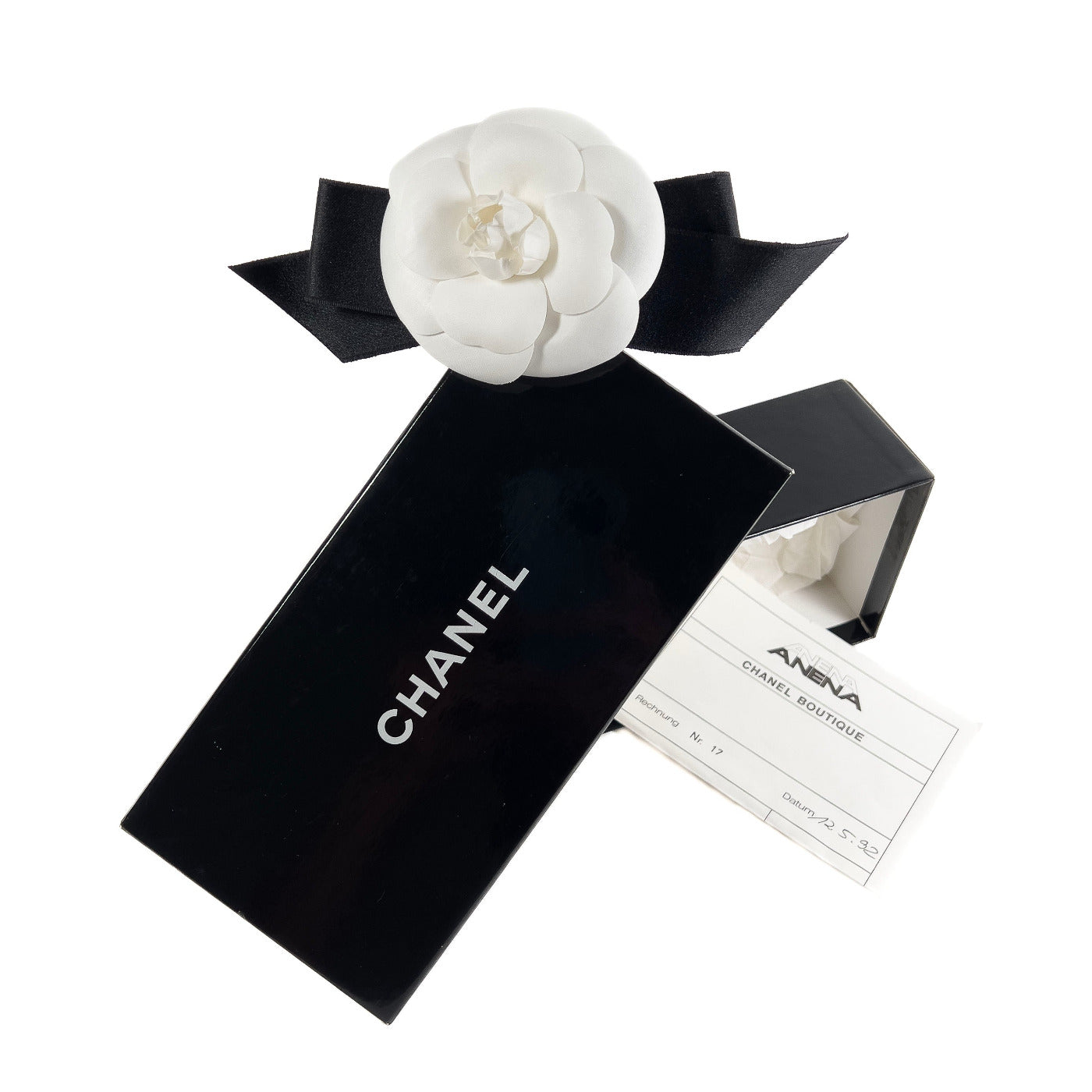 Vintage Chanel Brooch White Camelia on Black Ribbon.
A vintage Chanel brooch featuring a white camellia on a black ribbon, displayed alongside its original black packaging with the Chanel brand name and a boutique invoice.