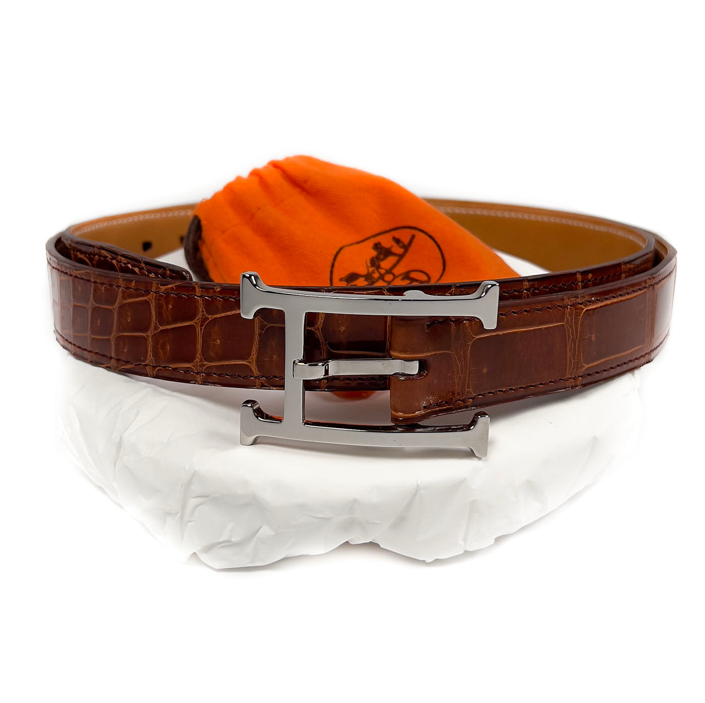 Pre-owned Hermès Cognac Porosus Crocodile Belt with Palladium Hardware Size 90, displayed on a white surface with an orange cloth.