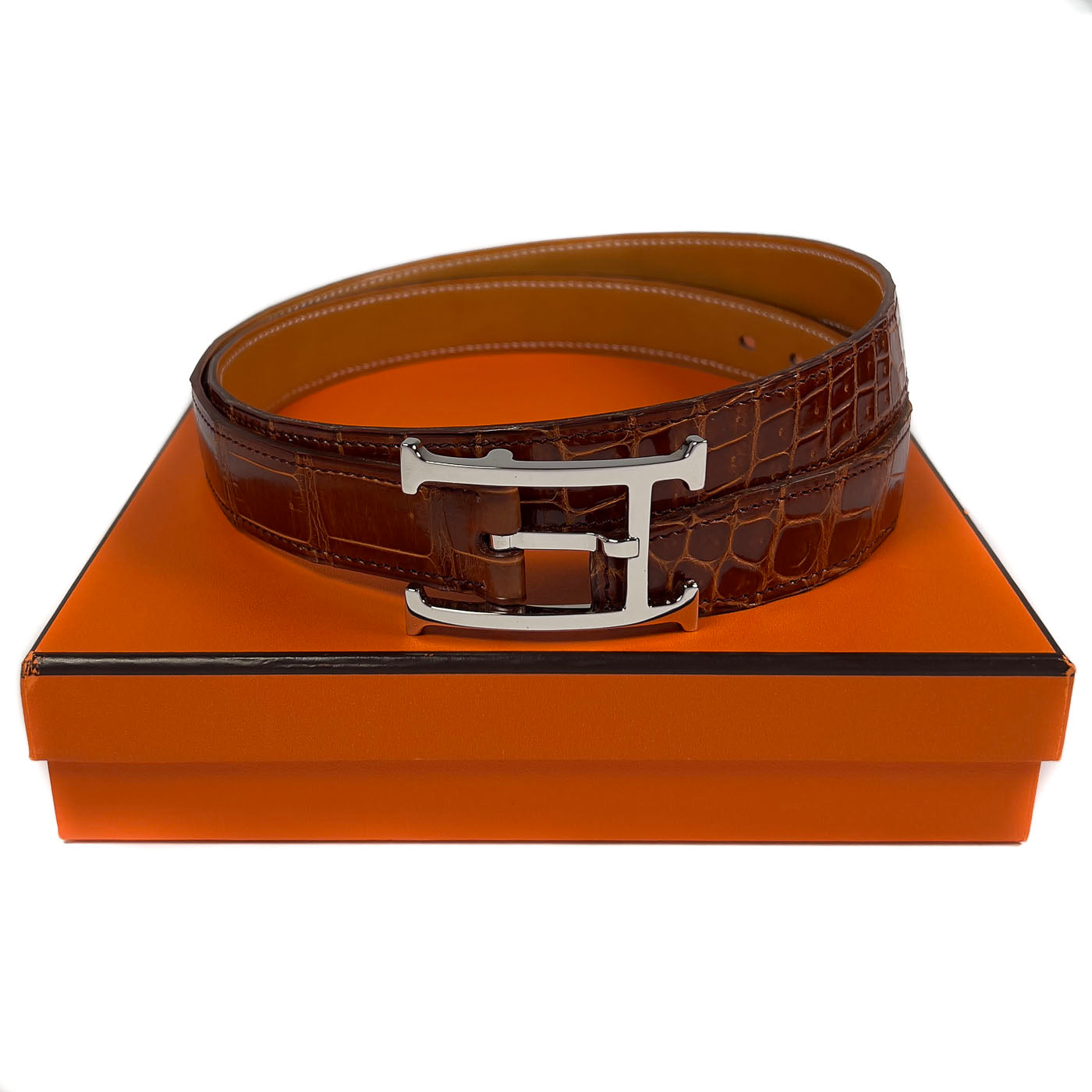 Pre-owned Hermès Cognac Porosus Crocodile Belt with Palladium Hardware Size 90, displayed on a white surface with an orange cloth.